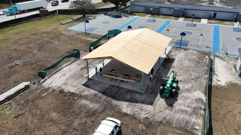 Drone view showing the progress of a new roof installation on an outdoor school shelter, featuring workers and a JCB telehandler.